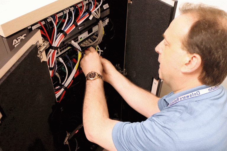 A person wearing a blue shirt and lanyard is working on wiring inside an APC-branded server rack. The person's detail-oriented focus is on the colorful bundle of cables they are managing. The cabinet doors of the server rack are open.