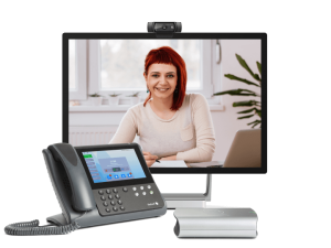 A woman with red hair is seen smiling on a computer monitor during a video call. In the foreground, there's a PT Mini 3020 conference phone with a display screen, alongside a small, silver speaker. The setup appears to be for remote communication or online meetings.