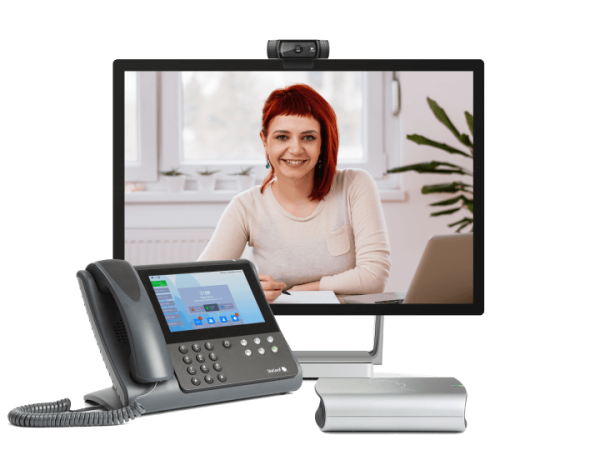 A woman with red hair is seen smiling on a computer monitor during a video call. In the foreground, there's a PT Mini 3020 conference phone with a display screen, alongside a small, silver speaker. The setup appears to be for remote communication or online meetings.