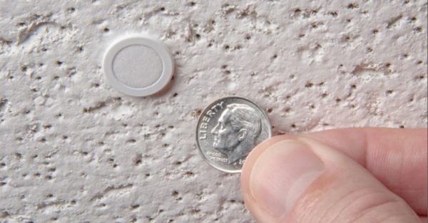 A hand holding a dime next to the CTG Audio CM-01 Ceiling Microphone - Silver pressed against a textured, off-white surface. The dime, depicting Franklin D. Roosevelt, provides a sense of scale for the microphone beside it.